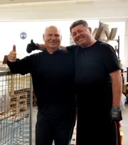 Zwei TEAMLOG Kollegen Arm in Arm in der Werkstatt, einer zeigt Daumen hoch